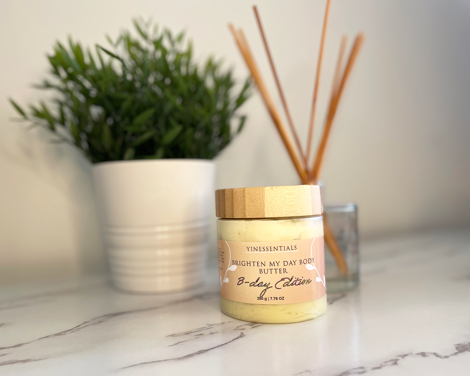 A jar of Brighten My Day Body Butter on a marble surface with a plant and reed diffuser in the background.