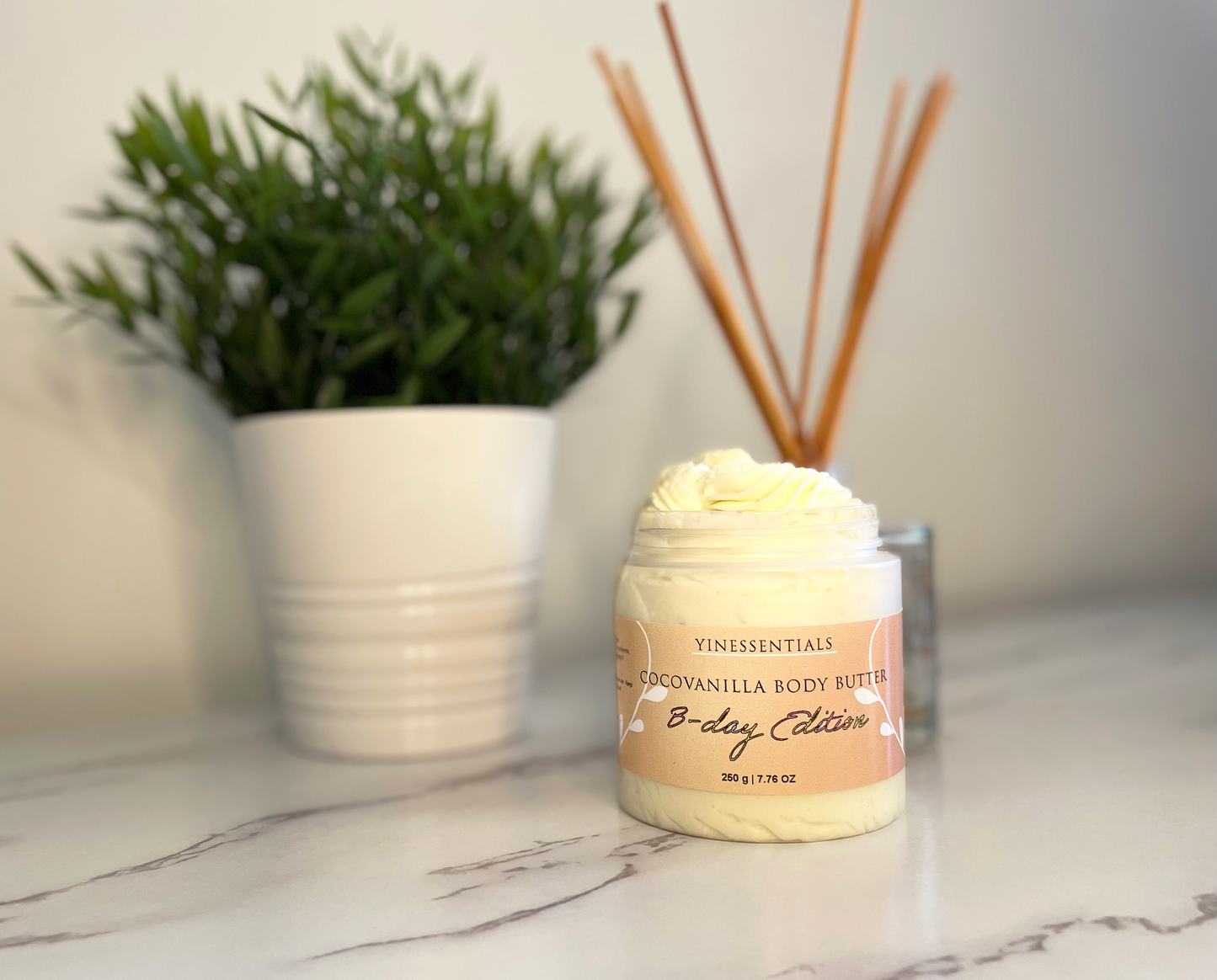 A jar of Cocovanilla Body Butter placed on a marble surface with a plant and some reed diffusers in the background.
