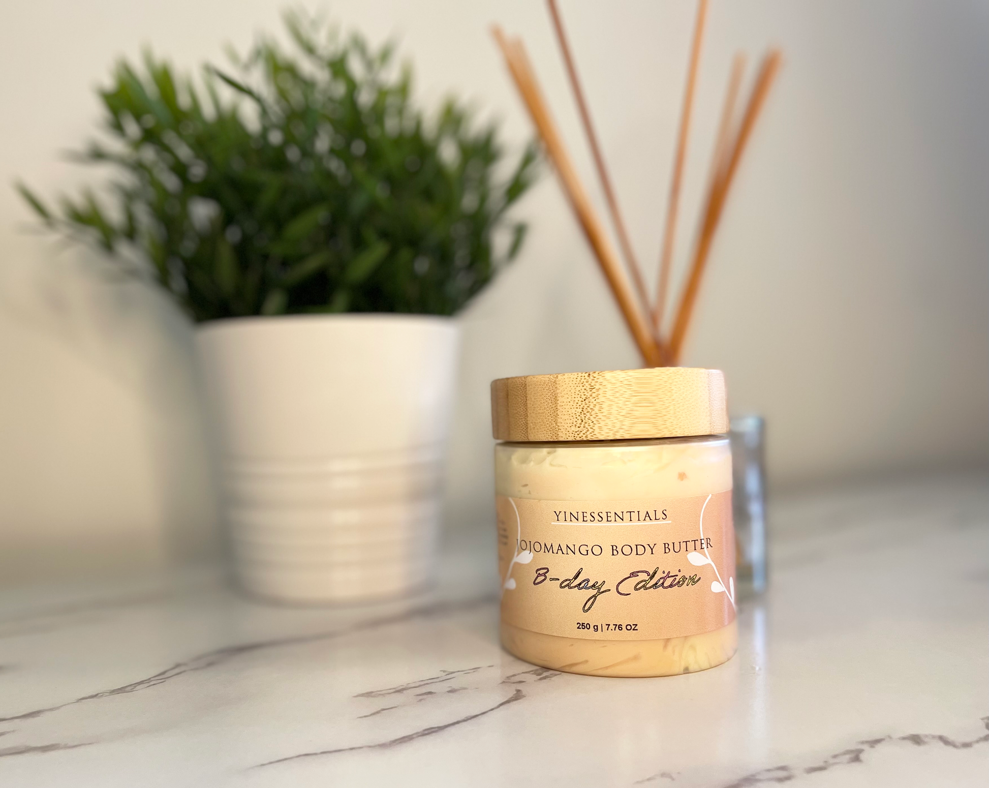 A jar of Jojomango Body Butter on a kitchen countertop with a plant and wooden reed diffusers in the background.